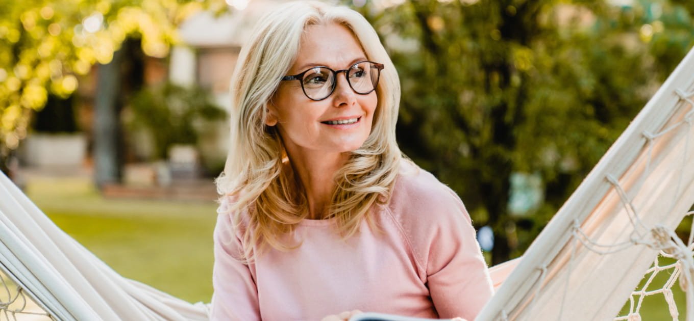 Dojrzała kobieta uśmiechnięta na hamaku z książką – objawy menopauzy u kobiet