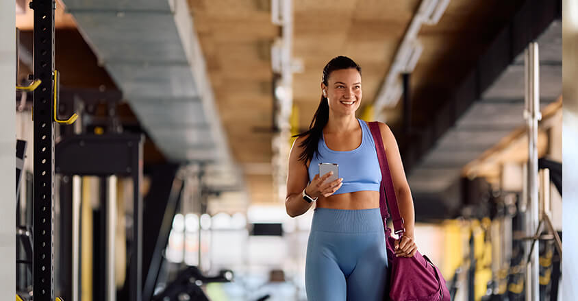 Młoda kobieta w siłowni z telefonem w ręku i torbą na ramieniu idzie na trening