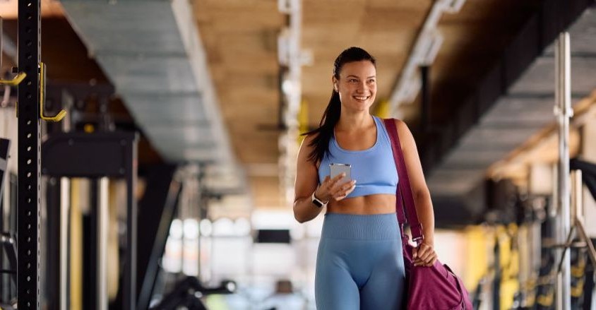 Młoda kobieta w siłowni z telefonem w ręku i torbą na ramieniu idzie na trening