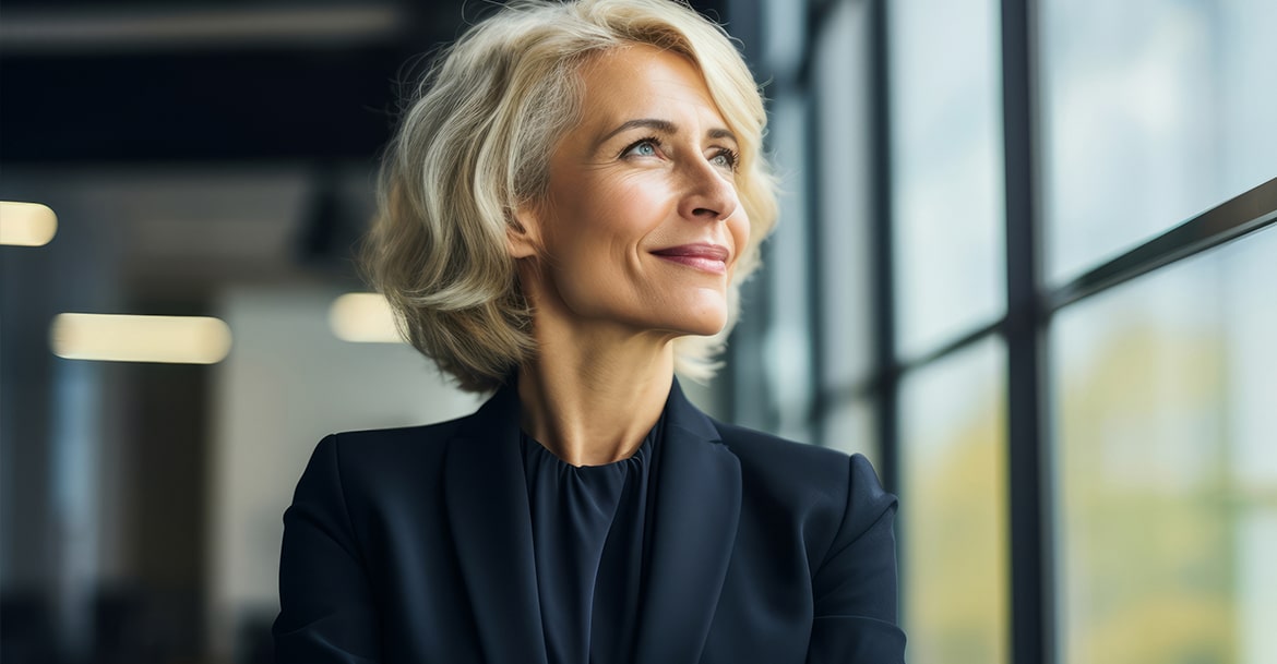 Piękna dojrzała kobieta w czarnym garniturze patrzy w okno i się uśmiecha - pierwsze objawy menopauzy