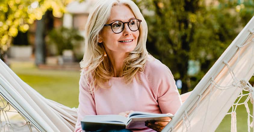 Dojrzała kobieta uśmiechnięta na hamaku z książką – objawy menopauzy u kobiet.