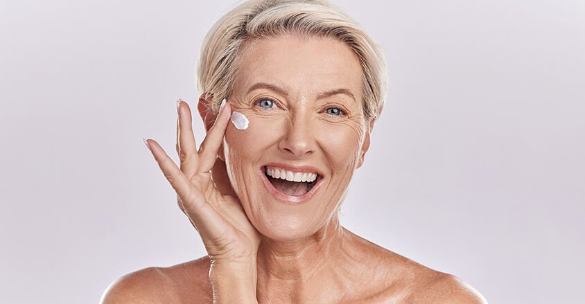 Roześmiana kobieta w dojrzałym wieku aplikująca krem na zmarszczki mimiczne